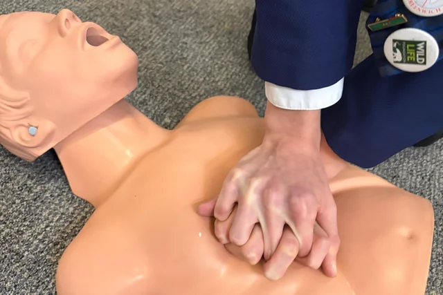 a pair of hands pressed onto the chest of a resuscitation manikin, a navy school blazer sleeve at the edge of the frame