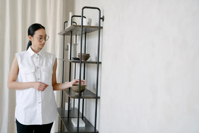 Woman styling a shelving unit with ceramics and decorative objects against a white wall