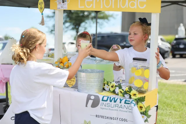 two girls at an outdoor stand passing a cup of lemonade from one pair of hands to another