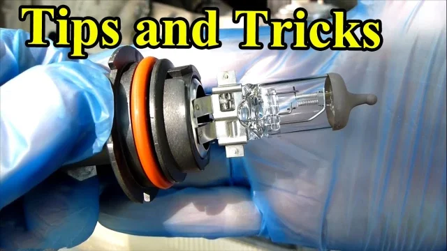 Gloved hands holding a halogen headlight bulb beside a headlight housing