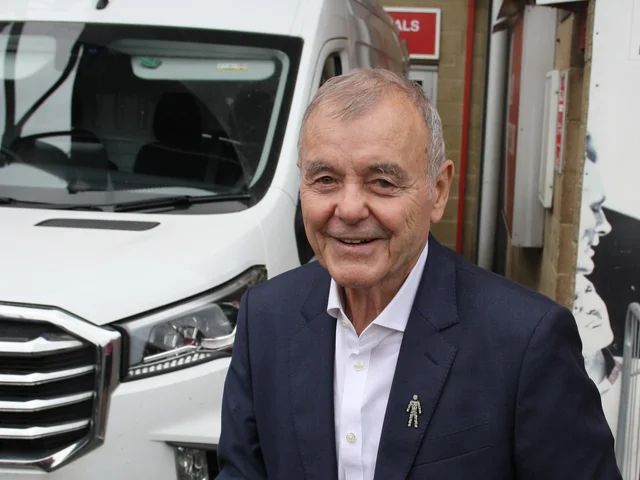 "A smiling older man in a dark suit and white open-necked shirt, standing outdoors in front of a white van"