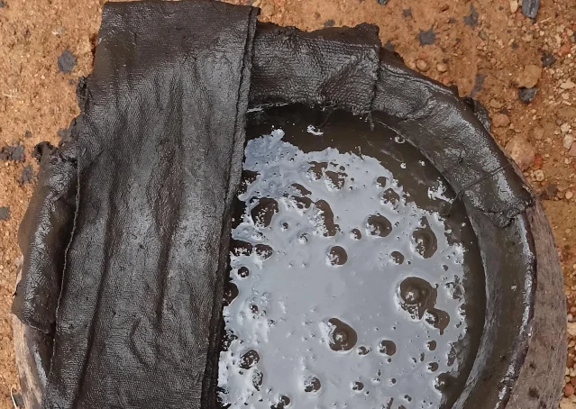 A dark textile resting in a shallow basin of muddy liquid, with reddish earth visible around it