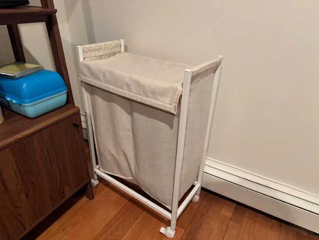 ENHET laundry bin with white metal frame and beige linen-like fabric bag on wheels, photographed in a bedroom corner next to a shelving unit