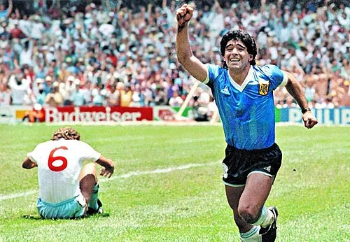 Maradona in Argentina's sky-blue jersey sprinting toward camera in celebration, an England player in white crouching on the grass behind him as Azteca's stands fill the background