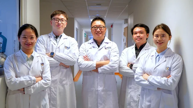 Five NUS researchers in white lab coats standing together in a corridor, arms crossed, facing the camera