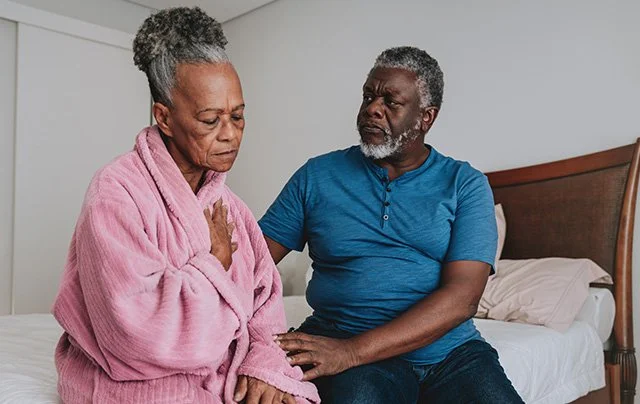 An elderly couple sitting on a bed; the woman holds her chest looking distressed while the man comforts her, illustrating the cardiovascular risk theme of the EDS and prolonged sleep latency study