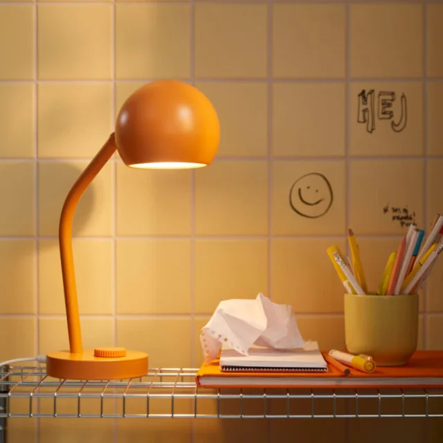 KORVSNÖRE orange LED desk lamp on a wire-shelf desk, dial dimmer visible on the base, lit against yellow tile wall