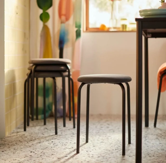 GULTARP stackable stools in a sunlit room with terrazzo flooring — three stools stacked near a wall, one pulled out to a dining table height