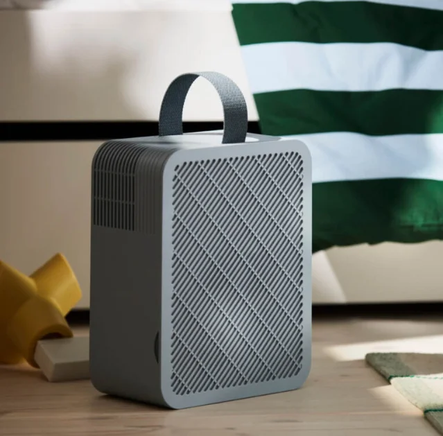 UPPÅTVIND portable air purifier — grey square unit with fabric textile carry handle, sitting on a wood floor beside a bed with striped cushions