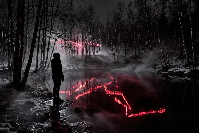 Woman's silhouette standing at the edge of a dark reflective forest pool, neon-red light threading through bare winter birch trees, mist low across the ground