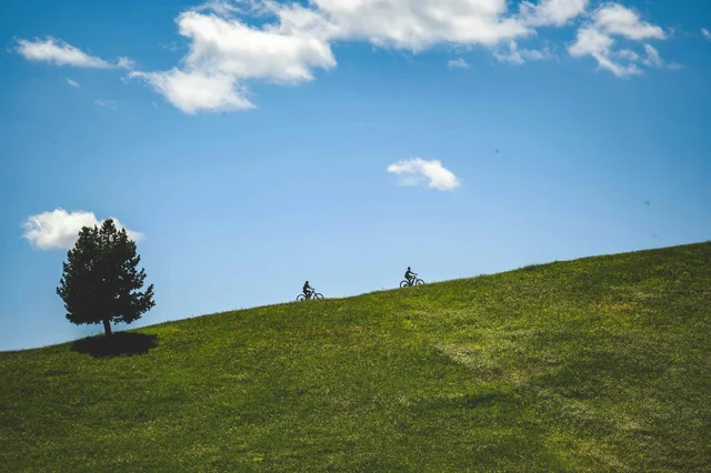 Two cyclists climbing a grassy hill under blue sky — terrain elevation is one of the biggest range variables