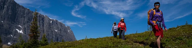 Three hikers in bright gear walking single-file along a green hillside; a sheer dark cliff wall rises on the left; blue sky above