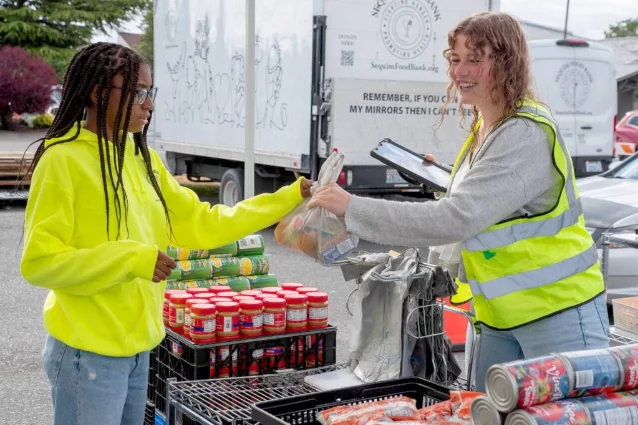 A teen volunteer hands food to another volunteer during distribution