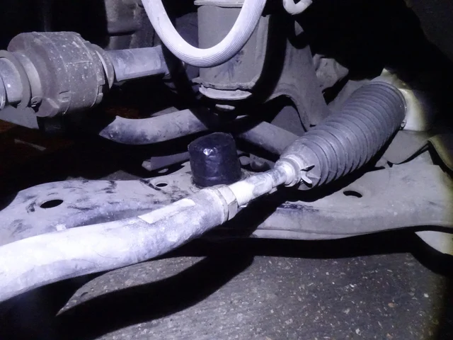 Underside of a car's front suspension: a black tape-wrapped cylindrical object lodged between the control arm and steering rack