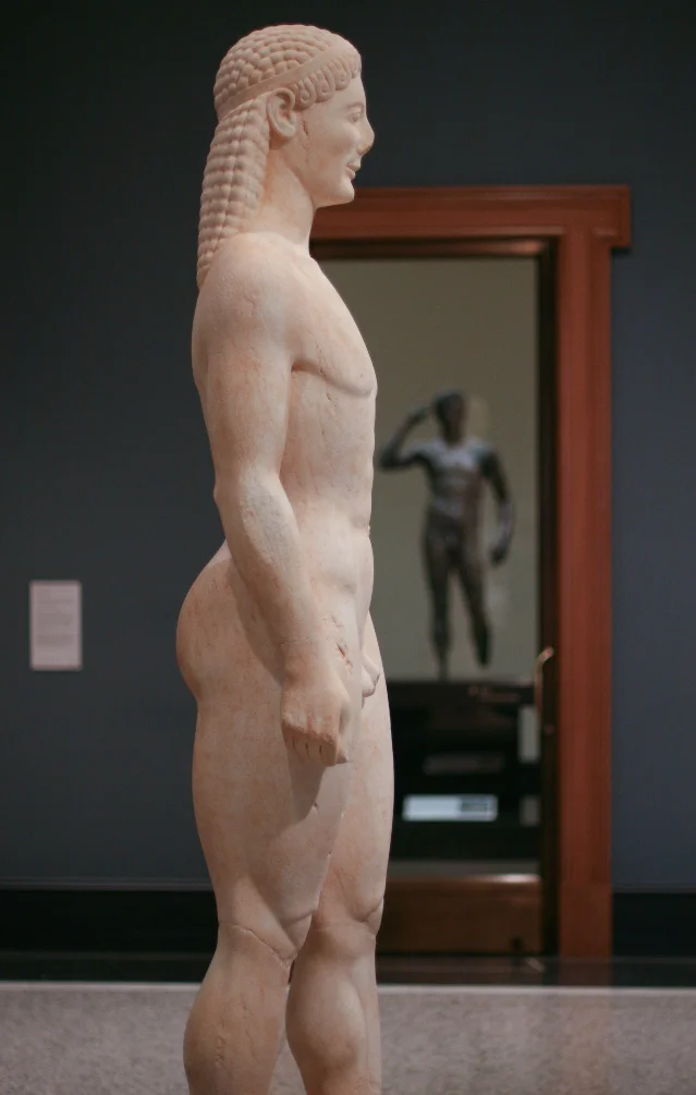 Getty Kouros (85.AA.40) seen from the side at the Getty Villa, with the Victorious Youth bronze visible through the doorway beyond