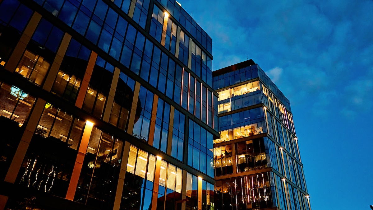 Two modern glass office buildings at twilight, windows lit with warm yellow interior light against a dark blue sky