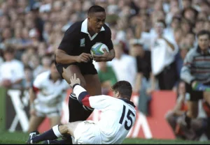 Jonah Lomu in All Blacks black jersey running hard past an England defender, ball in hand, 1995 Rugby World Cup