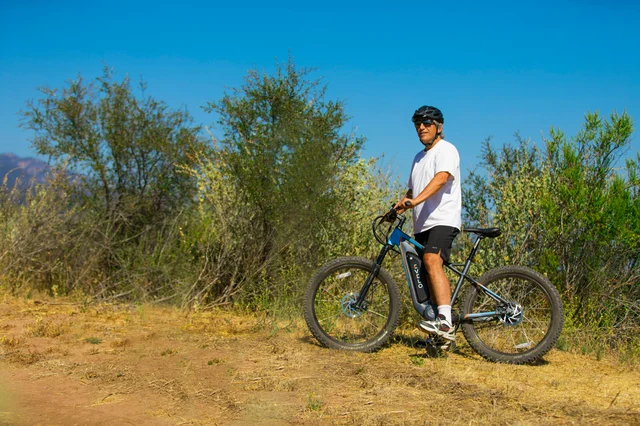 Man with helmet and sunglasses on an eBike on a dusty outdoor trail — real riding conditions vary significantly from flat-road estimates