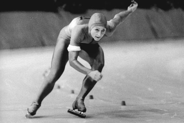 A black-and-white press photograph of a woman in a speed-skating suit and cap, leaning low with one arm swung back, skating between marker cones on an ice rink