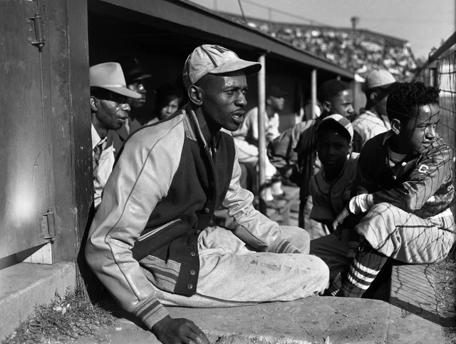 A black-and-white press photograph of a man in a varsity-style baseball jacket and pants sitting on the dugout step among players and bat boys