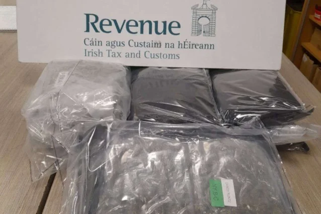 Five dark vacuum-sealed packages of varying sizes stacked on a wooden surface beneath a Revenue — Irish Tax and Customs sign