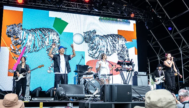 Leenalchi performing at WOMAD festival in 2023, the full seven-piece on stage against a backdrop showing illustrated tigers
