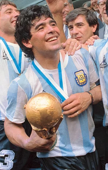 Maradona in Argentina's blue-and-white striped kit holding the 1986 FIFA World Cup trophy aloft, medal ribbon around his neck, teammates celebrating behind him
