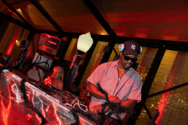 DJ performing under red lights at a Late Night Editions event, NY Yankees cap and sunglasses, mixing board and speakers visible, gold sequined backdrop
