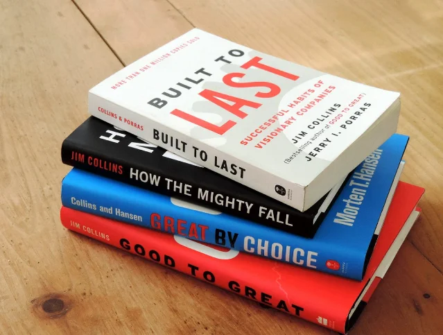 A stack of four Jim Collins books — Built to Last, How the Mighty Fall, Great by Choice, Good to Great — on a wooden table