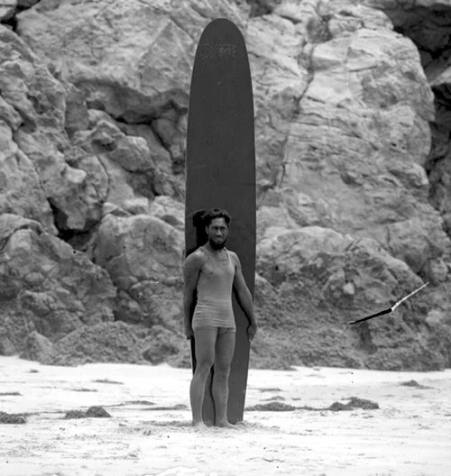 Duke Kahanamoku standing on a beach at Corona Del Mar, California, leaning against his massive solid redwood surfboard, c. 1920