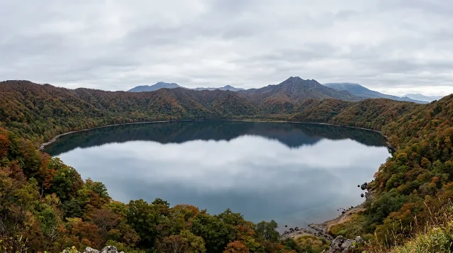 Volcanic caldera lake surrounded by forested hills in autumn — still, dark water reflecting an overcast sky, mountains visible in the distance, no people, viewed from a ridge