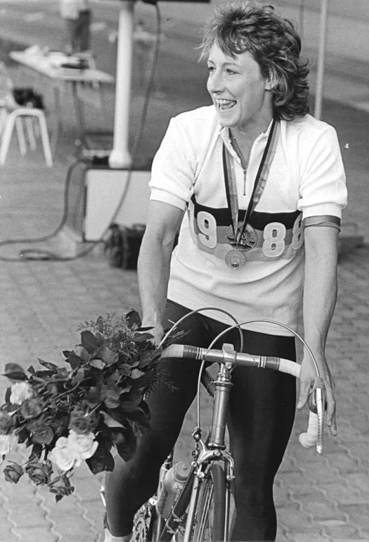 A black-and-white press photograph of a woman in a white short-sleeved jersey and dark leggings standing behind a track bicycle, a large bunch of roses across the handlebars and a medal hanging at her chest