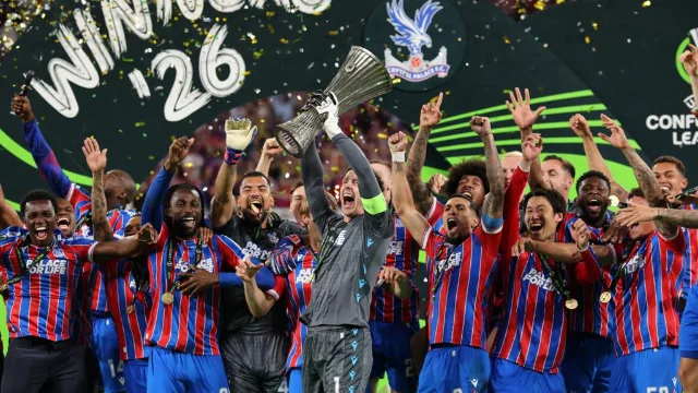 Crystal Palace squad celebrating with the Conference League trophy, gold confetti, red-and-blue kits, 'WINNERS 26' branding