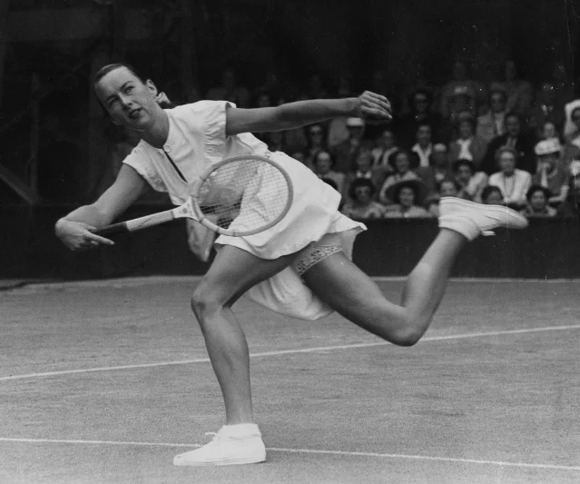 Gussie Moran in the controversial Teddy Tinling-designed white dress, lace-trimmed knickers visible as she reaches wide on Court One, Wimbledon 1949, packed spectator stands behind her