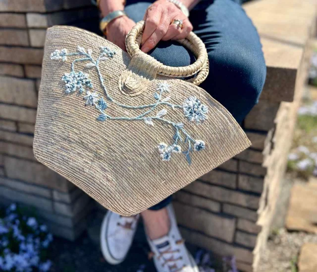 A hand holding a natural straw handbag with hand-embroidered blue floral sprigs, photographed against a brick wall in natural sunlight