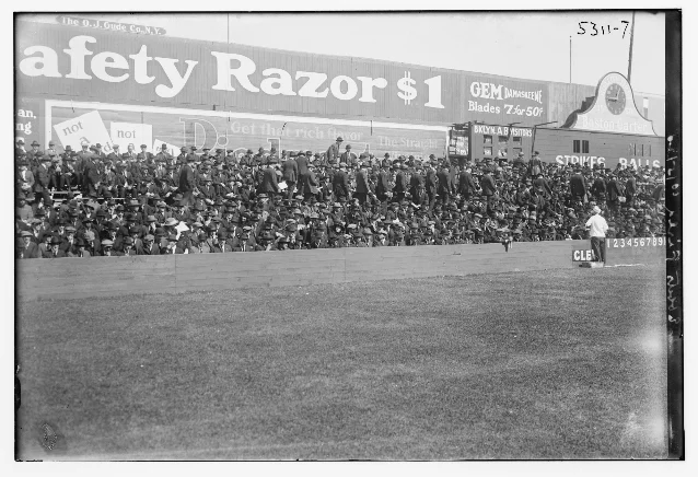 A packed grandstand at Ebbets Field seen from field level behind a low wooden outfield fence, with advertising boards along the wall and a scoreboard on the right