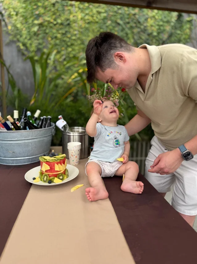Sam Altman with his child at a birthday celebration, fruit cake made from watermelon and kiwi on the table, green plant wall background