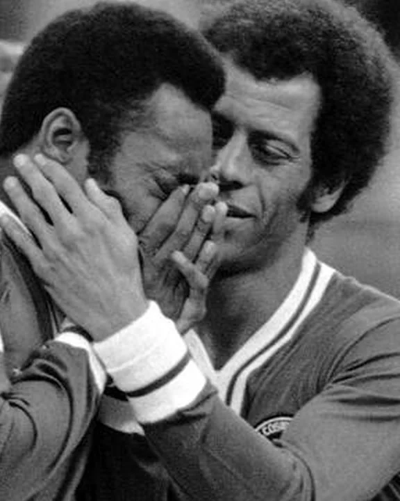 A man in a football shirt covers his face with his hand while a teammate in the same kit puts an arm around his shoulder in a stadium