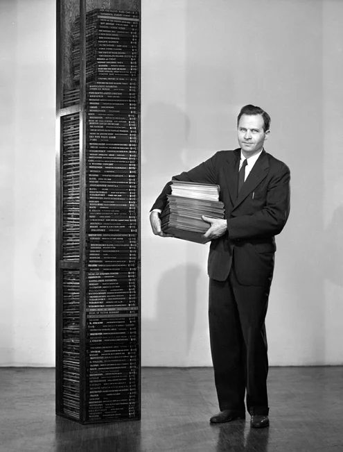 Peter Goldmark standing beside a tower of 78 rpm records while holding a stack of LPs, demonstrating the LP's storage advantage