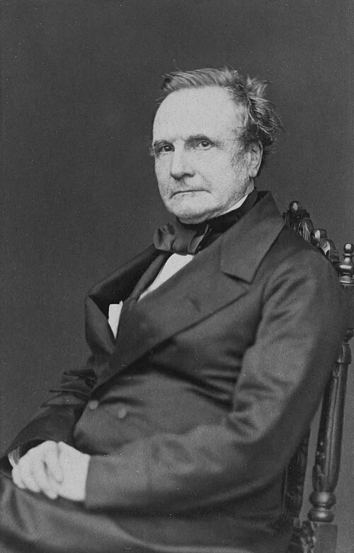 Charles Babbage, photographed circa 1860, seated in a chair wearing a dark coat, looking directly at the camera with a composed expression