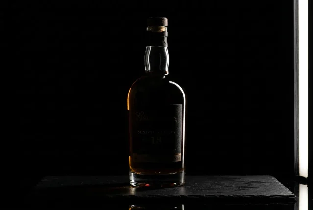 Strip softbox positioned at the right side of a dark studio — raking edge light creates a precise vertical highlight down the right face of a whiskey bottle, deep shadow on the left