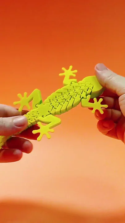 Neon yellow-green 3D printed articulated gecko held between two hands against an orange background, showing the segmented body being gently flexed