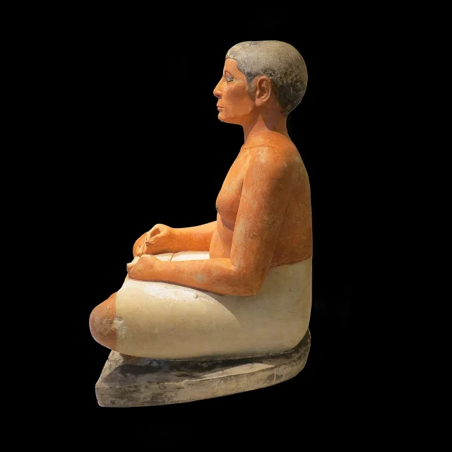 Side view of the Seated Scribe, showing the cross-legged pose, rounded abdomen, white kilt, and integrated base
