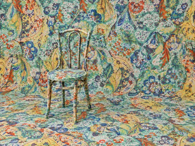 A studio scene in which a colourful floral print covers a wooden chair, the floor and the backdrop in one continuous field