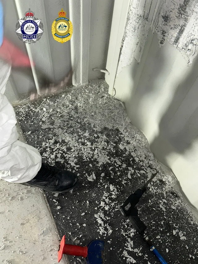 AFP officer in white protective suit standing next to the corner of the deconstructed shipping container; white paint flakes scraped off the walls cover the floor; AFP and ABF badges visible top-left