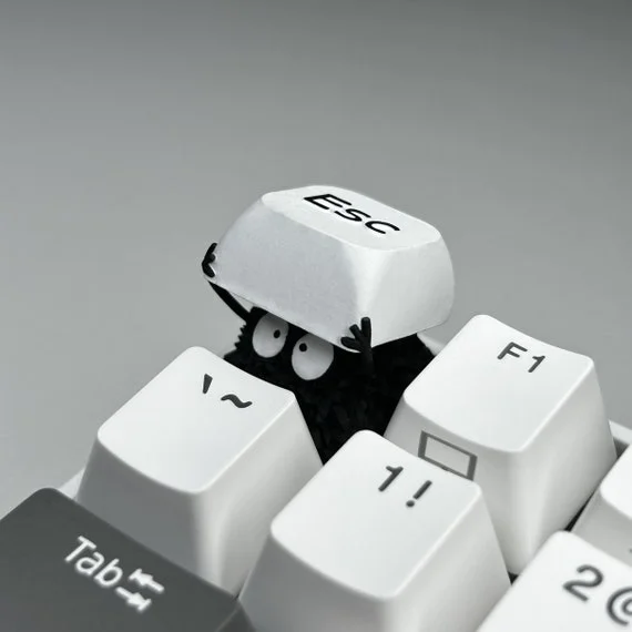 The "Keyboard Gremlin" artisan keycap by CobaltPrints — a black dust-creature figure peeking out from between keyboard keys while holding the ESC keycap overhead, macro photography on white keys