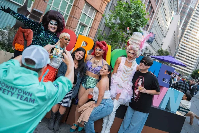 Outdoor crowd at Drag Me Downtown event at 101 California Plaza — drag performers and attendees in a sun-lit Financial District plaza, colorful props and city buildings in the background