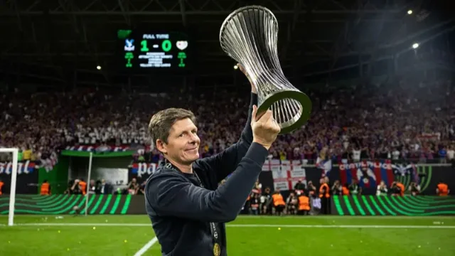 Oliver Glasner holding the Conference League trophy aloft, stadium scoreboard showing Crystal Palace 1-0 behind him
