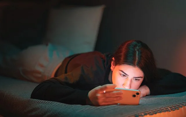 A young woman lying in bed in a darkened room, holding a phone close to her face lit by the screen's glow