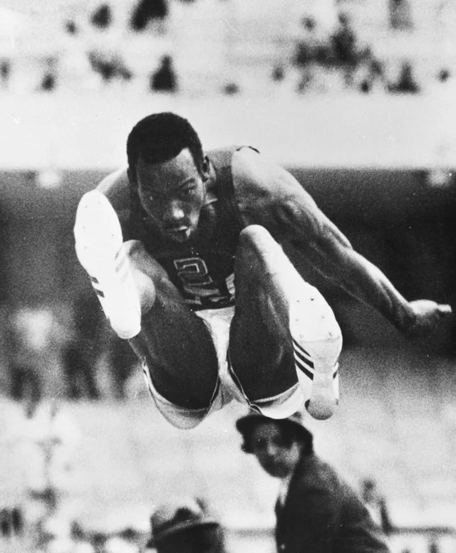 Bob Beamon in mid-air above the long jump pit, knees drawn up, arms out, a blurred crowd behind him